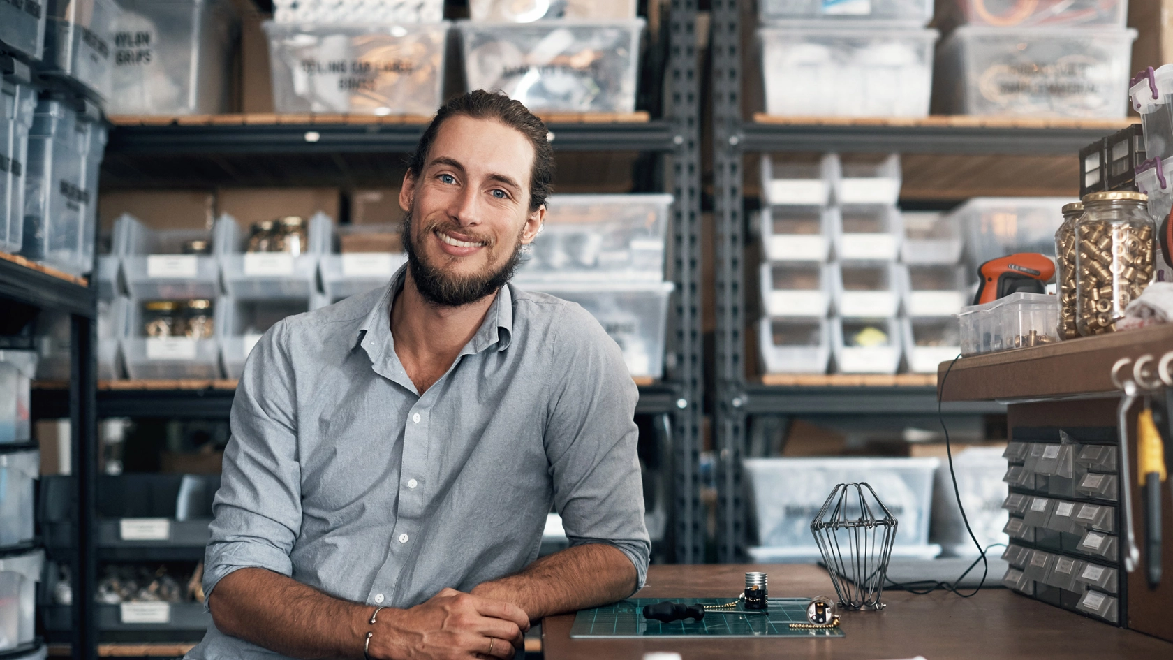 Junger Mann mit Hemd leht mit dem Arm auf einer Arbeitsfläche vor einem Regallager mit kleineren Metallteilen und blickt direkt in die Kamera