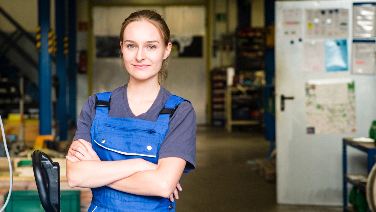 Eine junge Frau in einer blauen Arbeitshose und einem blauen T-Shirt steht mit verschränkten Armen da und schaut freundlich