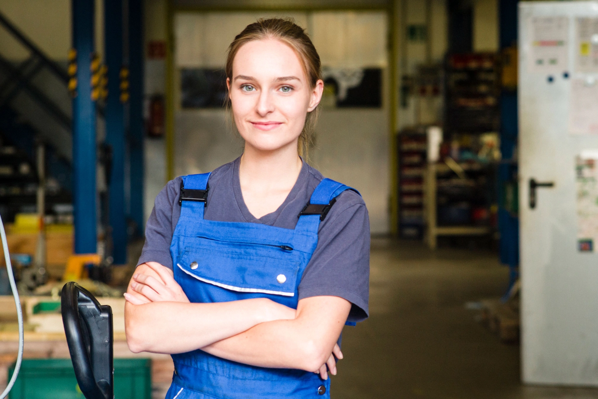 Eine junge Frau in einer blauen Arbeitshose und einem blauen T-Shirt steht mit verschränkten Armen da und schaut freundlich